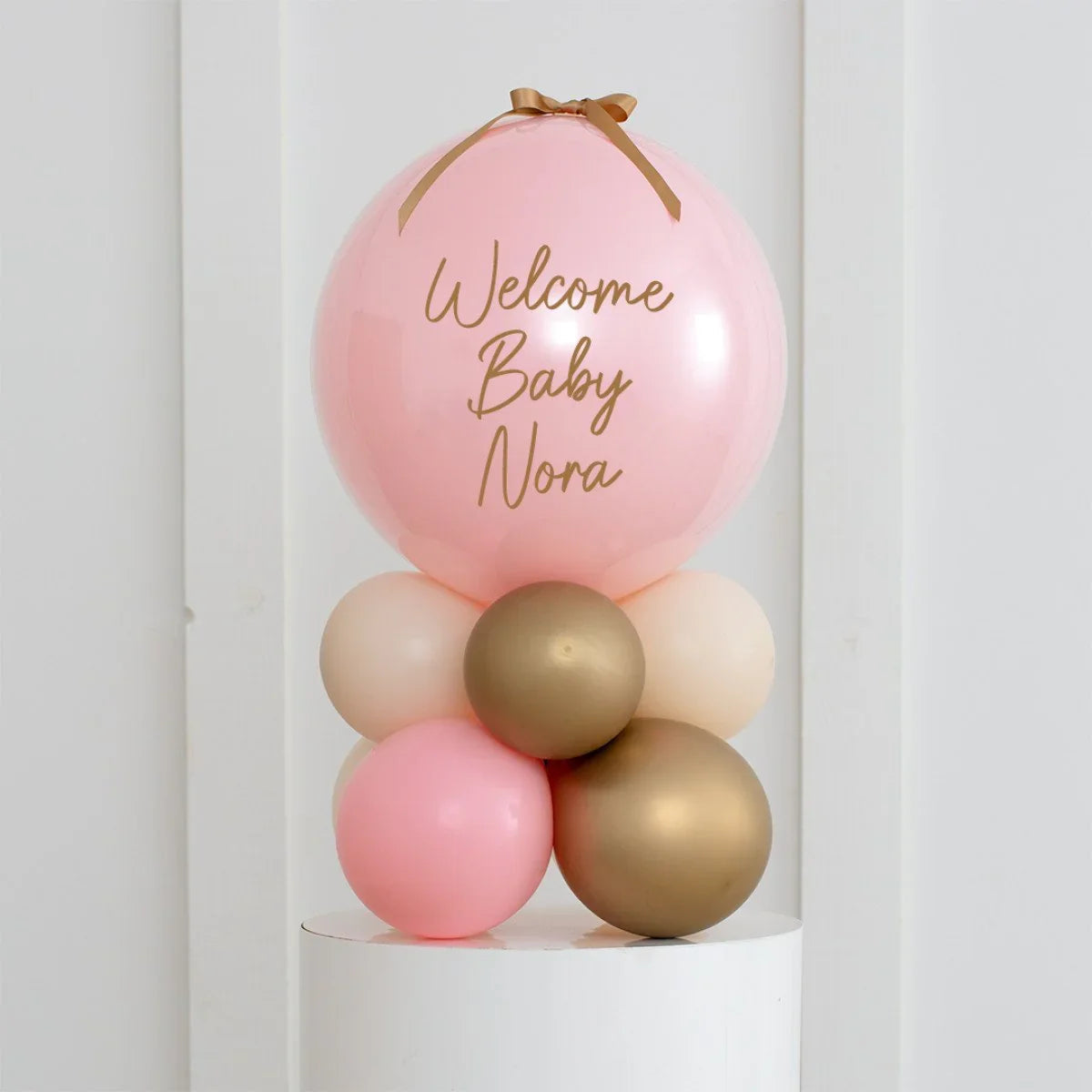 Pink and gold baby shower balloon arrangement with Welcome Baby Nora text and ribbon